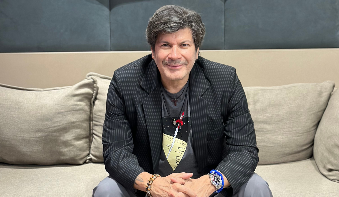 Em novembro ocorrerá o show de Paulo Ricardo em Curitiba celebrando os 40 anos de carreira com nova turnê. Na imagem, vemos o artista, um homem branco de cabelo preto, usando um blazer preto, camisa e calça cinzas. O artista está sentado em um sofá, com as mãos entrelaçadas sobre os joelhos e olhando para a câmera.
