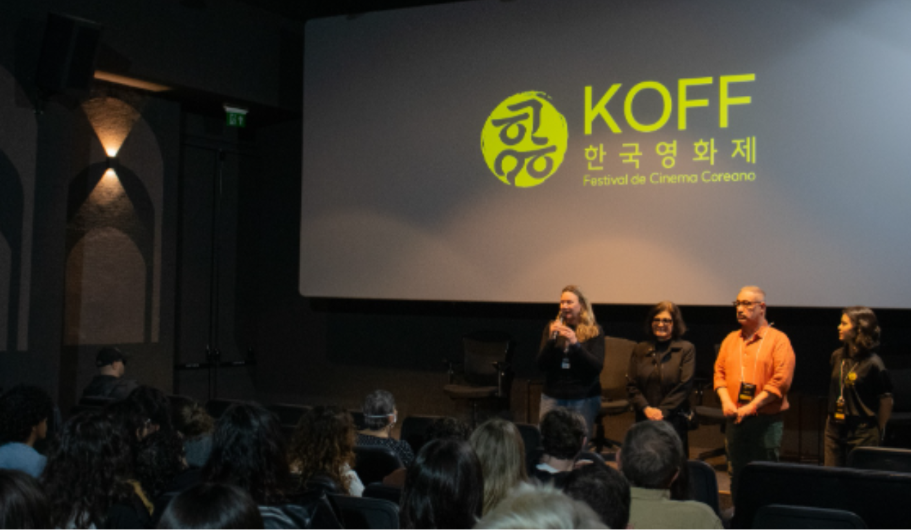 Um festival de filmes coreanos estará em Curitiba em setembro. Na imagem, vemos uma exibição em Porto Alegre. A exibição ocorre em uma sala escura, com um telão exibindo a logo da Koff. Próximo à câmera vemos cabeças de pessoas que estavam no assistindo ao festival, mais distante da câmera vemos quatro pessoas em pé. Da esquerda para a direita: a primeira é uma mulher branca e loira, que usa uma blusa preta e calça jeans azul, ela segura um microfone em suas mãos; a segunda é uma mulher branca de cabelo castanho, que usa um óculos de armação preta, blusa e calça pretas; o terceiro é um homem branco de cabelos grisalhos, com um óculos de armação preta, vestindo uma camisa laranja e calça jeans; a quarta é uma mulher negra de cabelo preto, vestindo camiseta e calça pretas, com um crachá com a logo do evento.