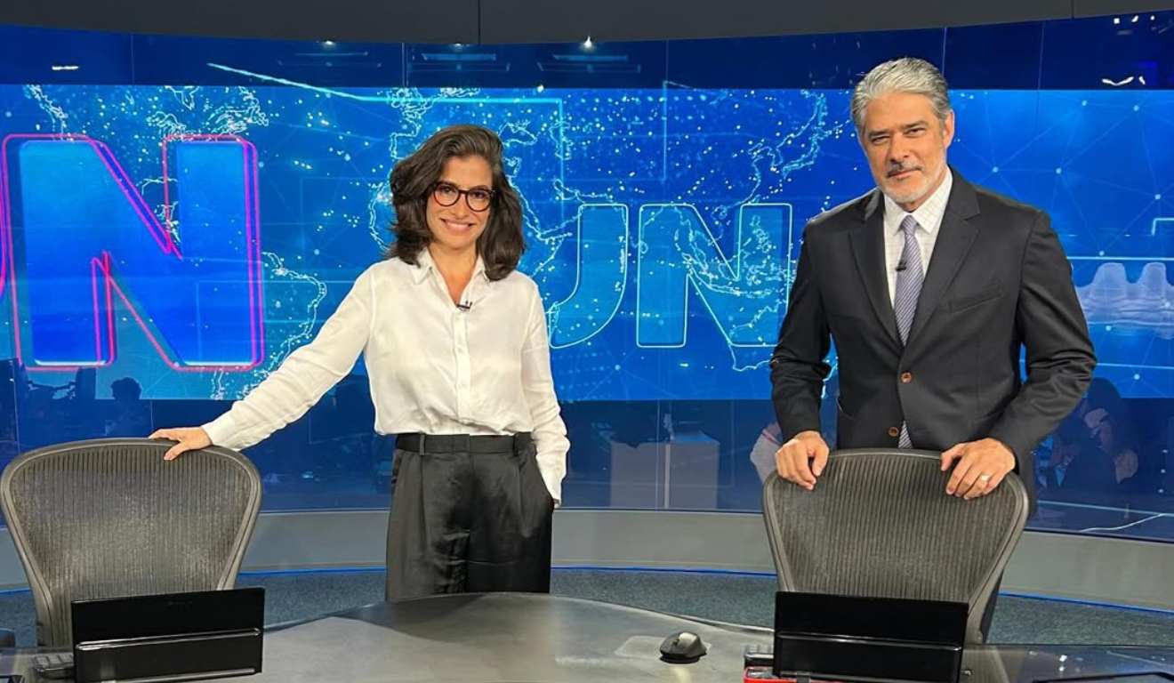 William Bonner anunciou que está saindo do Jornal Nacional. Na imagem, vemos ele e Renata Vasconcellos, na bancada do programa. Renata, uma mulher branca de cabelo preto, usa um óculos de armação preta, uma camisa branca e uma calça preta. Ela está com a mão direita apoiada na cadeira e a esquerda no bolso da calça. Ao seu lado direito está William Bonner, um homem branco, de cabelos grisalhos e barba branca. Ele usa um terno azul escuro, uma camisa branca e uma gravata azul com listras brancas.