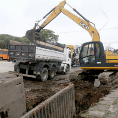Um cruzamento será bloqueado por obras do Inter 2 em Curitiba. Na imagem, vemos uma escavadeira colocando terra em um caminhão. Um trabalhador, utilizando EPI, está ao lado.