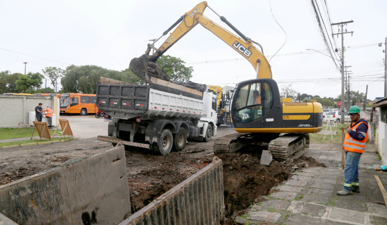Um cruzamento será bloqueado por obras do Inter 2 em Curitiba. Na imagem, vemos uma escavadeira colocando terra em um caminhão. Um trabalhador, utilizando EPI, está ao lado.