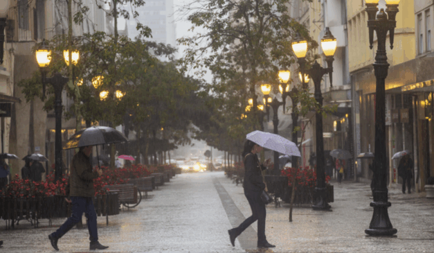 Curitiba registrou chuva em quase metade do ano. Na imagem, vemos a Rua das Flores, em Curitiba, durante um dia de chuva. Duas pessoas estão andando e segurando guarda-chuvas.
