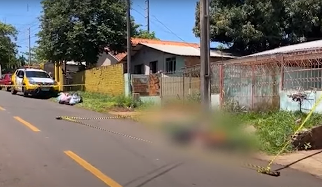 Um homem, de 33 anos, foi morto a tiros enquanto andava de bicileta. Na imagem, vemos a cena do crime, isolada pela Polícia Militar. Uma viatura da PM está presente atrás do corpo, que está borrado.