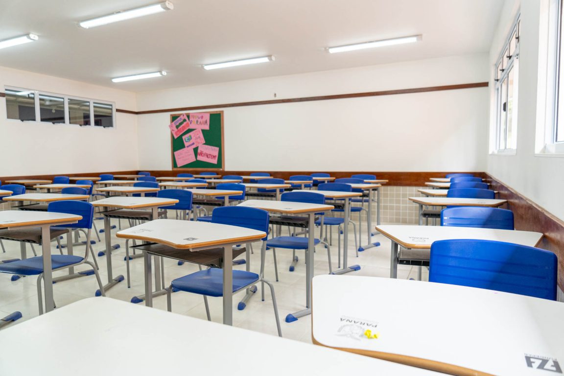 Imagem com sala de aula vazia com várias cadeiras e quadro de professor