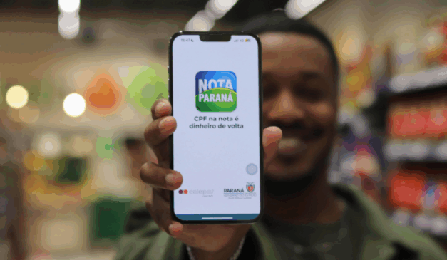 Nesta quinta-feira (9), será realizado o sorteio do Nota Paraná de outubro. Na imagem, vemos um celular com o aplicativo do Nota Paraná aberto. Um homem negro está segurando o celular mas está ao fundo da imagem, desfocado.