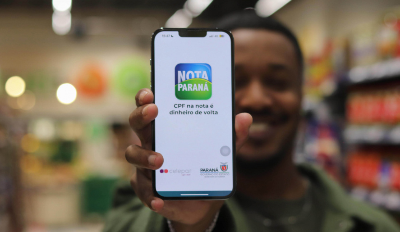 Nesta quinta-feira (9), será realizado o sorteio do Nota Paraná de outubro. Na imagem, vemos um celular com o aplicativo do Nota Paraná aberto. Um homem negro está segurando o celular mas está ao fundo da imagem, desfocado.