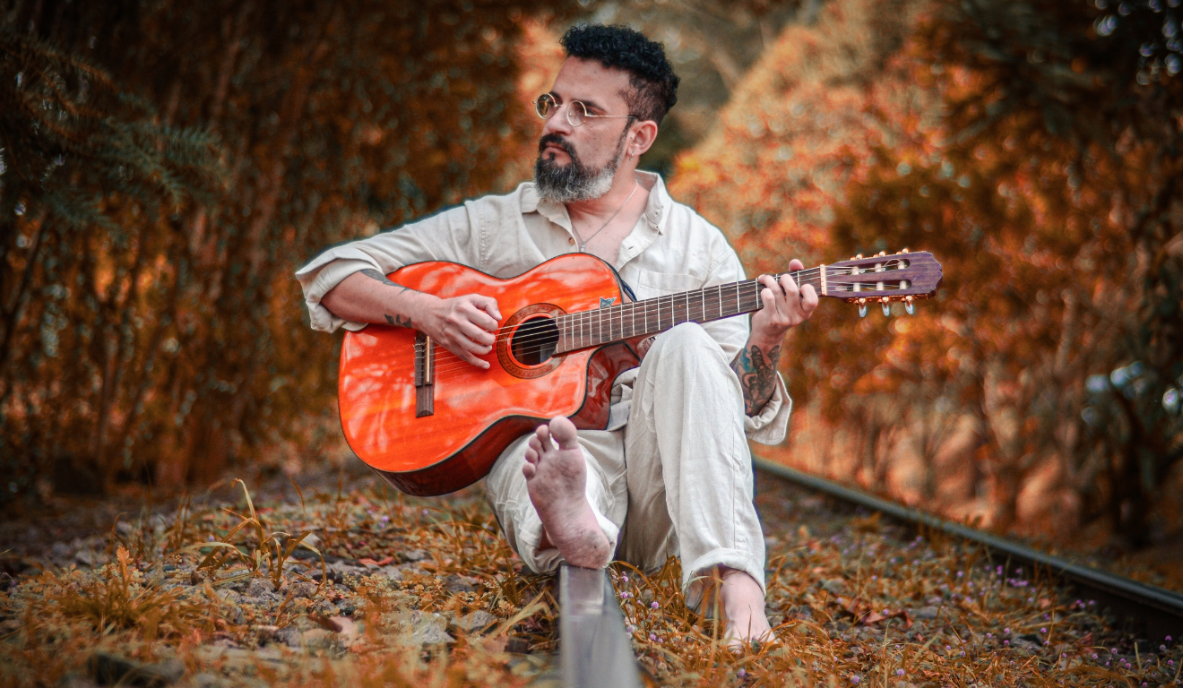 cantor Dabliu, com violão na mão, sentado no trilho do trem, em referência ao espetáculo Dabliu Canta Milton - Tudo Será Como Antes