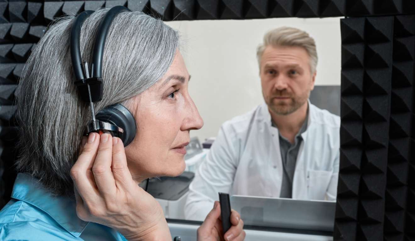 Idosa realizando exame de audiometria com médico em outra sala