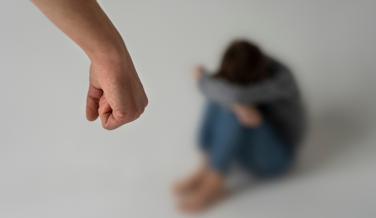 Homem ameaça de morte sua ex-companheira. Na iamgem ilustrativa, vemos uma mão branca em primeiro plano. Ao fundo, há uma mulher desfocada, ela está sentada, com os braços sobre os joelhos, escondendo o rosto nos braços.