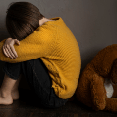 Um homem foi preso suspeito de estuprar as duas netas. Na imagem ilustrativa, vemos uma criança de costas para um urso de pelúcia. A criança, de camiseta amarela e calça preta, e está ajoelhada, com o rosto escondido nos braços, que estão sobre os joelhos.