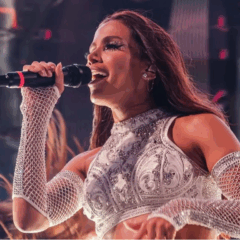 A pré-venda dos ingressos do Ensaios da Anitta em Curitiba esgotou em 20 minutos. Na imagem vemos Anitta, uma mulher para de cabelo preto, usando uma roupa cinza e segurando um microfone na mão direita, durante um show.