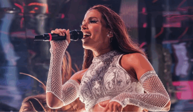 A pré-venda dos ingressos do Ensaios da Anitta em Curitiba esgotou em 20 minutos. Na imagem vemos Anitta, uma mulher para de cabelo preto, usando uma roupa cinza e segurando um microfone na mão direita, durante um show.