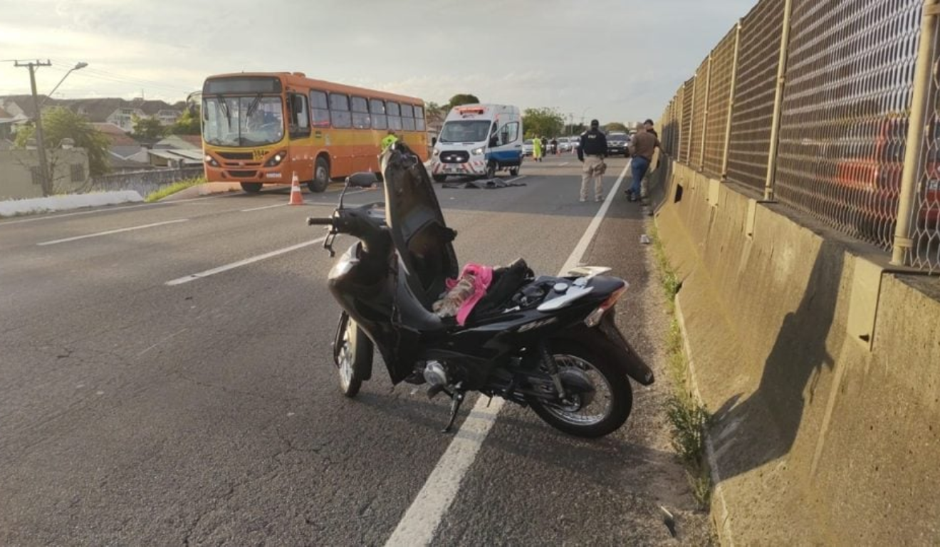 Jovem sofre acidente com motocicleta na BR-277, em Curitiba. Na imagem, vemos a motocicleta parada na rodovia, com uma ambulância e um ônibus ao fundo.