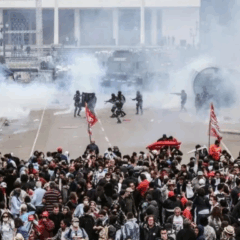 STF determina que Paraná indenize feridos no confronto do Centro Cívico em 2015. Na imagem, vemos, em primeiro plano, os manifestantes e, ao fundo, agentes policiais armados, com fumaça próxima a eles.