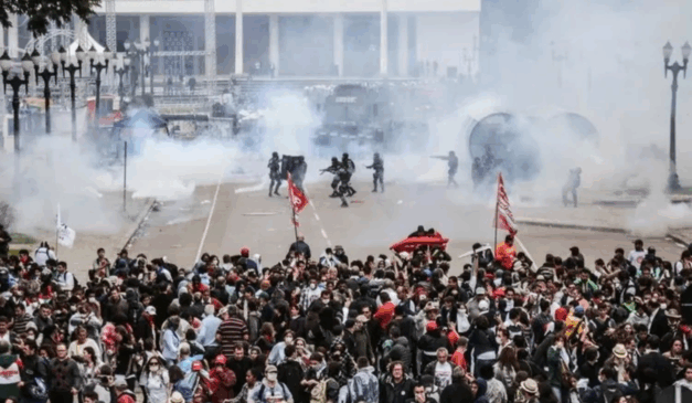 STF determina que Paraná indenize feridos no confronto do Centro Cívico em 2015. Na imagem, vemos, em primeiro plano, os manifestantes e, ao fundo, agentes policiais armados, com fumaça próxima a eles.