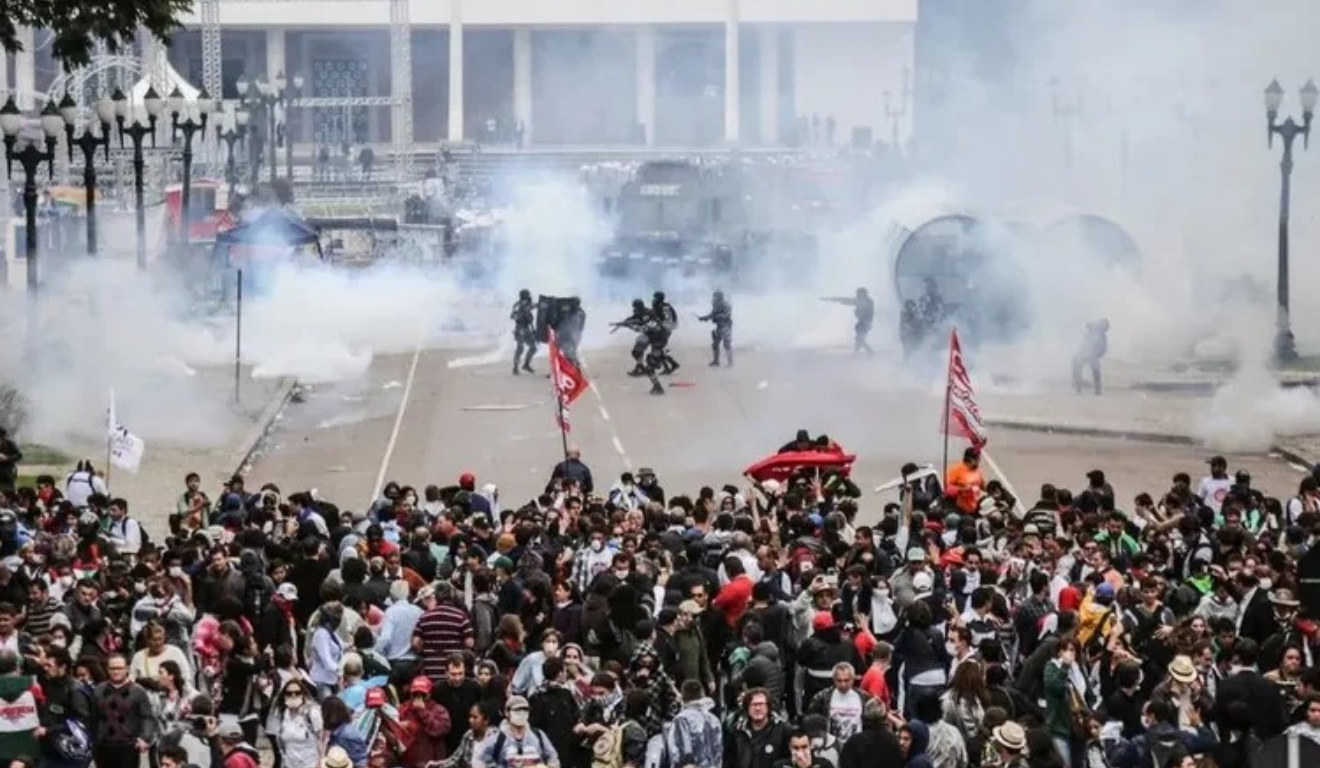 STF determina que Paraná indenize feridos no confronto do Centro Cívico em 2015. Na imagem, vemos, em primeiro plano, os manifestantes e, ao fundo, agentes policiais armados, com fumaça próxima a eles.