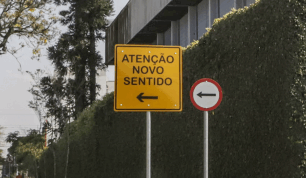 Quatro ruas de Curitiba terão alteração de sentido a partir do dia 14 de novembro. Na imagem, vemos uma placa amarela com o texto "Atenção Novo Sentido", indicando o sentido único de circulação de uma rua.