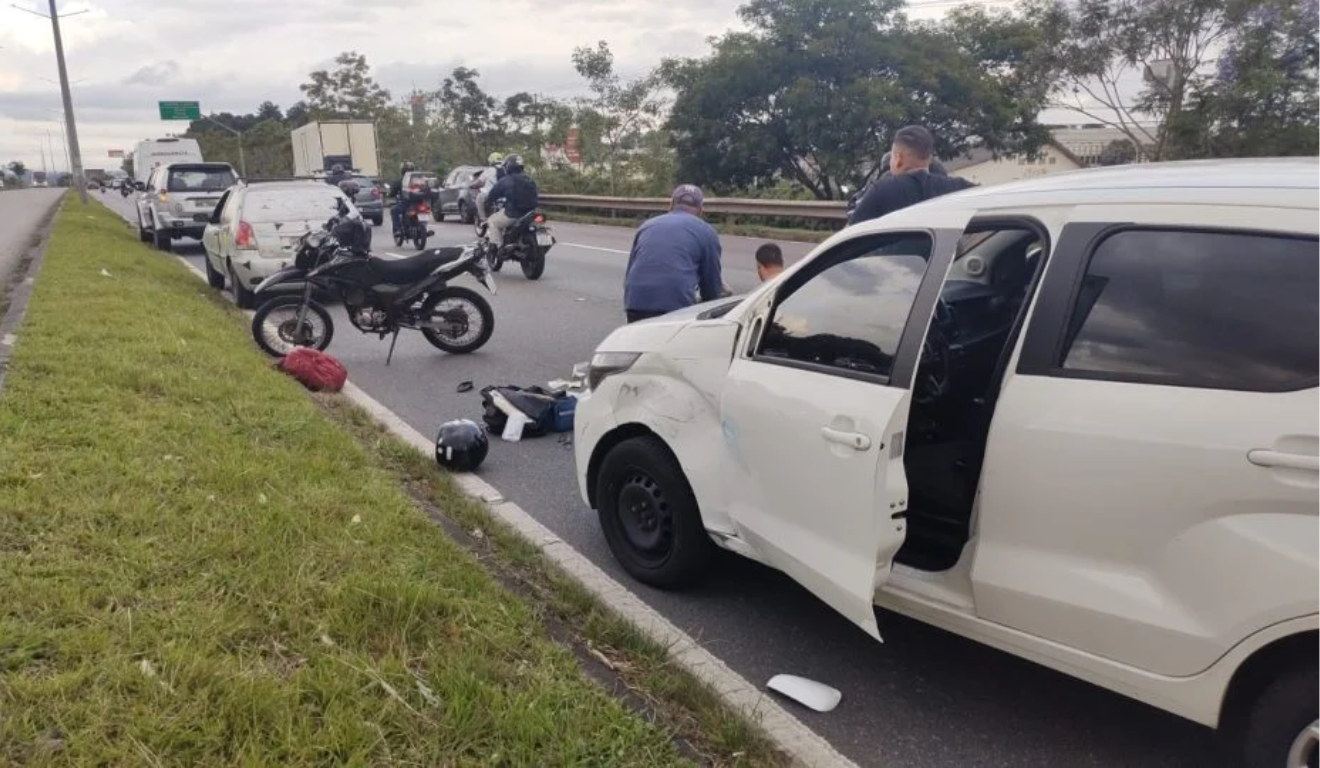 Cena do acidente na Linha Verde, em Curitiba, que envolveu três veículos. Um deles, o Fiat Uno branco, aparece em primeiro plano, com a moto da vítima logo em frente, no chão.