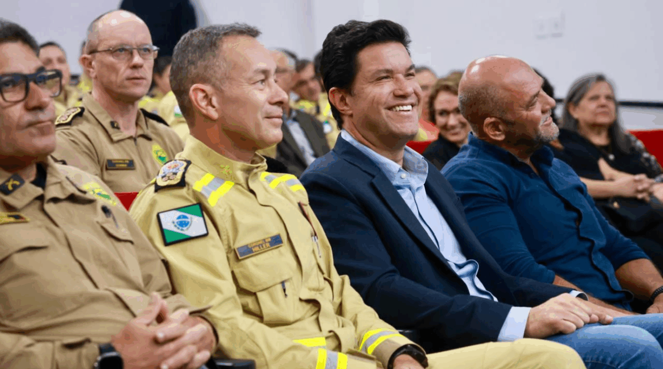 Guto Silva rindo, sentado ao lado do Coronel Hudson Teixeira e de comandantes do Corpo de Bombeiros do Paraná, durante homenagem que recebeu em Rio Bonito do Iguaçu