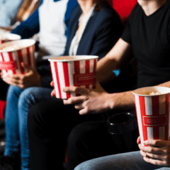 Na imagem ilustrativa, vemos pessoas sentadas em cadeiras de cinema, com pacotes de pipoca, representando eventos culturais que ampliarão o acesso à meia-entrada no Paraná.