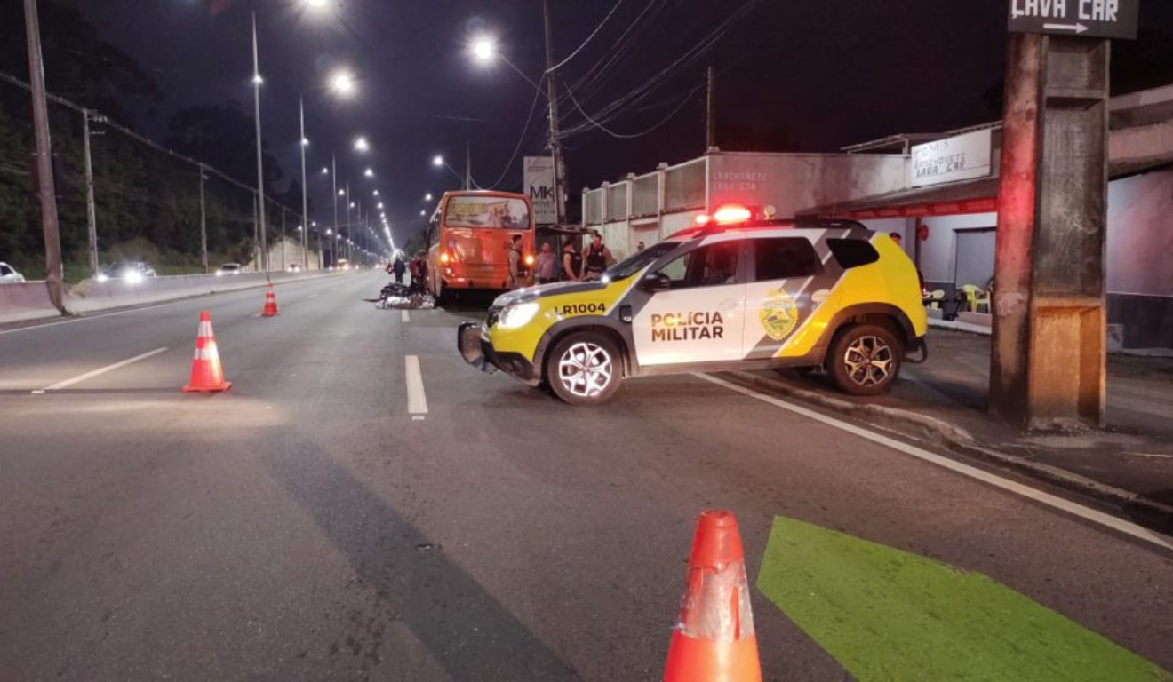 Na imagem, vemos o ônibus e a moto envolvidas no acidente. No primeiro plano, uma viatura da Polícia Militar; o motociclista bateu no ônibus, caiu e foi atropelado por um caminhão, em Piraquara.