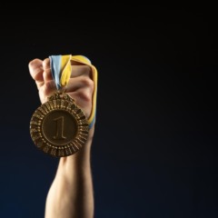 A imagem apresenta um plano m&eacute;dio focado no bra&ccedil;o e punho cerrado de um atleta, erguido verticalmente contra um fundo escuro e minimalista com um leve gradiente azulado na base. O Objeto Central: Uma medalha de ouro reluzente pendurada no punho, exibindo o n&uacute;mero "1" gravado no centro. A medalha possui bordas trabalhadas com padr&otilde;es geom&eacute;tricos e est&aacute; presa a uma fita de cores azul e amarela.