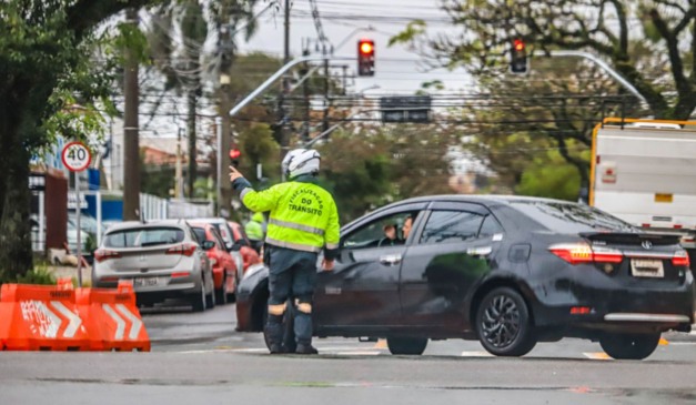 Guarda de trânsito de Curitiba orientando um carro que está passando por um trecho que tem uma rua bloqueada para obras.