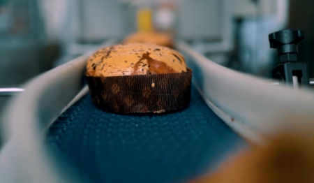Uma esteira industrial transporta panetones recém-assados em uma linha de produção. O foco principal está em um panetone com cobertura de chocolate e granulado, envolto em papel pardo tradicional. Uma esteira industrial transporta panetones recém-assados em uma linha de produção. O foco principal está em um panetone com cobertura de chocolate e granulado, envolto em papel pardo tradicional.